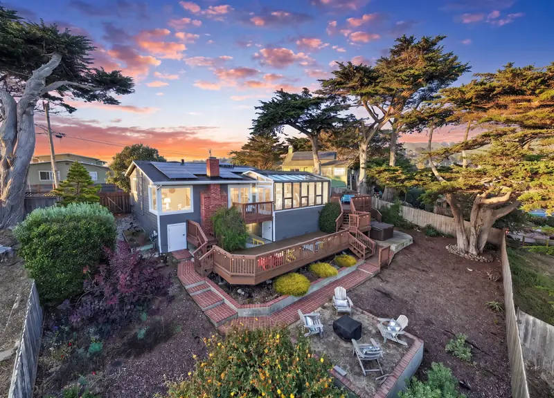 Drone view of a hillside home with a wraparound deck among cypress trees at dusk, Pacifica