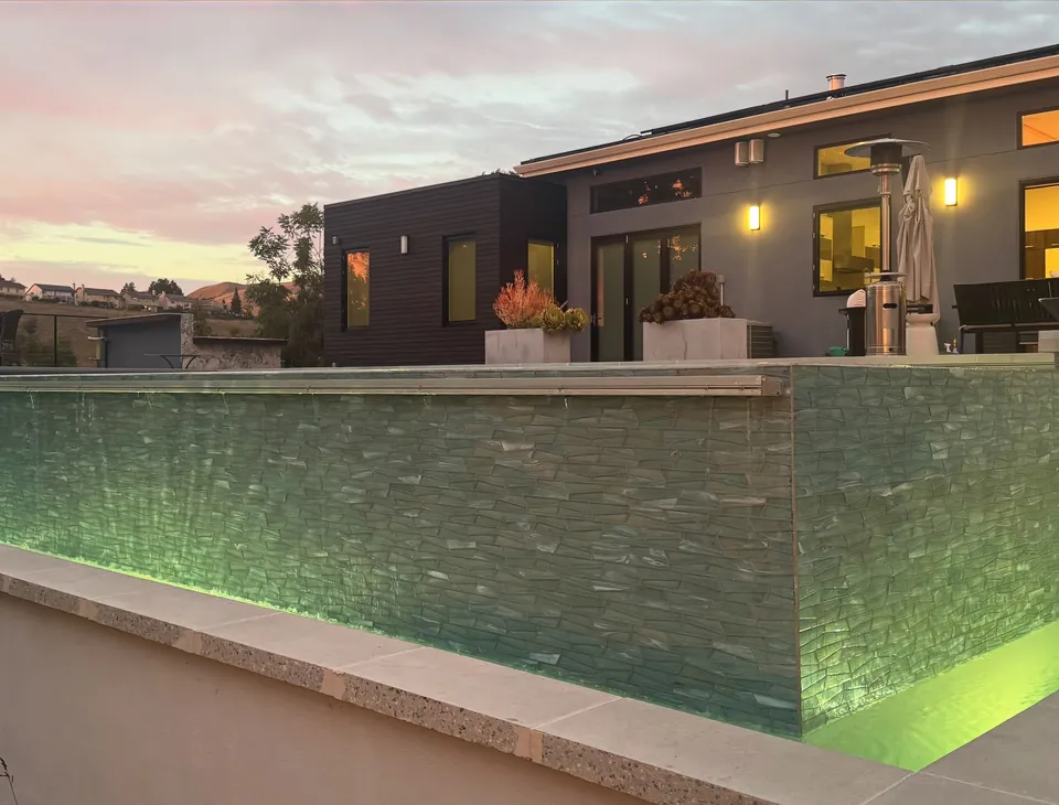Corner of the raised glass-tile pool wall with the catch basin lit green below a modern home at dusk