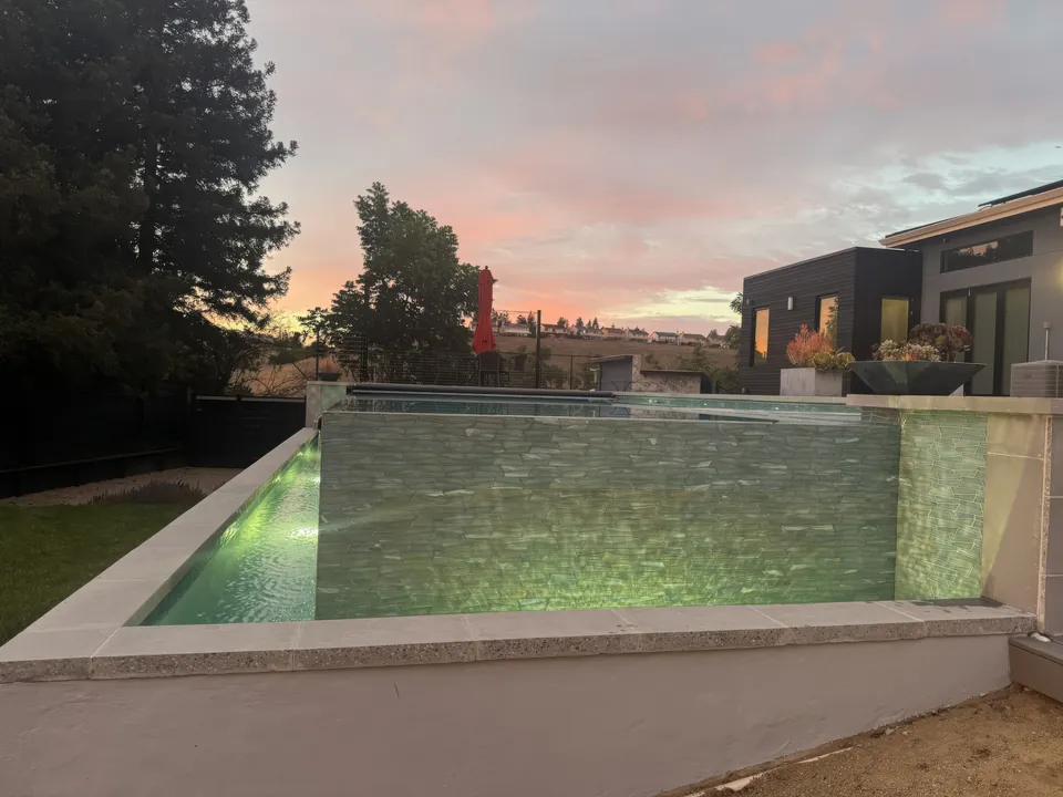 Raised vanishing-edge pool with its glass-tile wall and catch basin glowing under a pink sunset