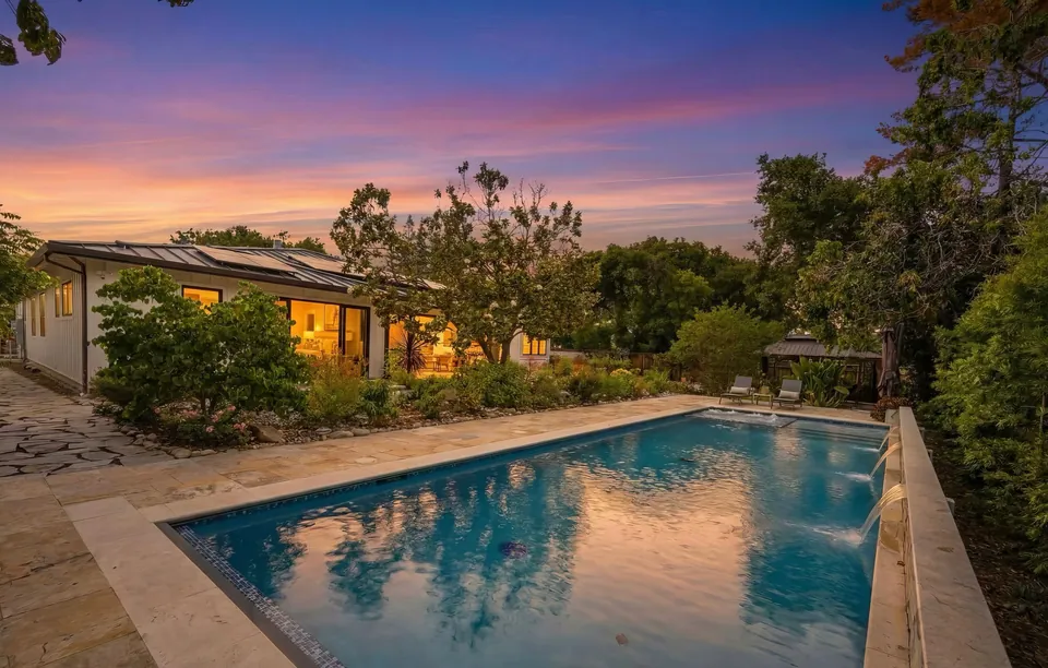 Rectangular pool with sheer descents beside a lit single-story home at sunset